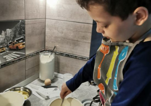 Chłopiec w kuchennym fartuszku podczas mieszania łyżką ciasta w kuchni.